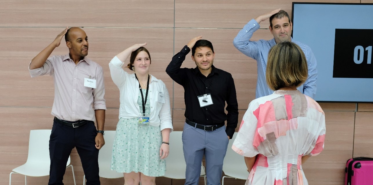 "4 people standing in a row, patting their heads. One person in the foreground with their back to us, telling them to do so."