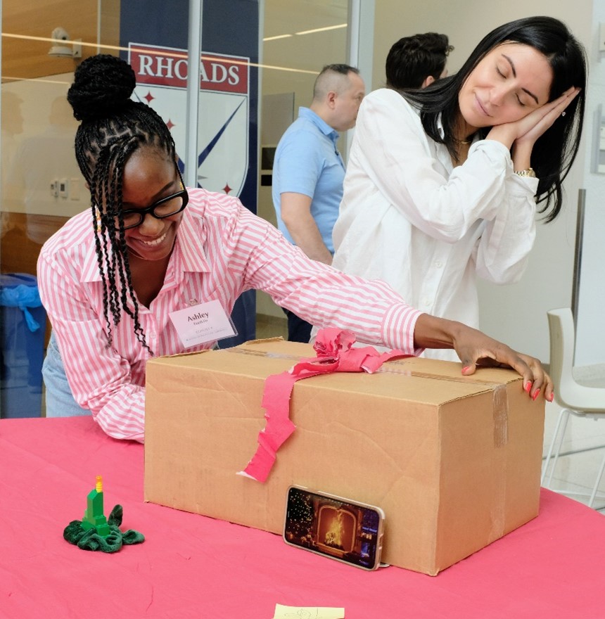 "One person placing a box on a table while a second person pantomimes sleep. The box has a pink ribbon on top."
